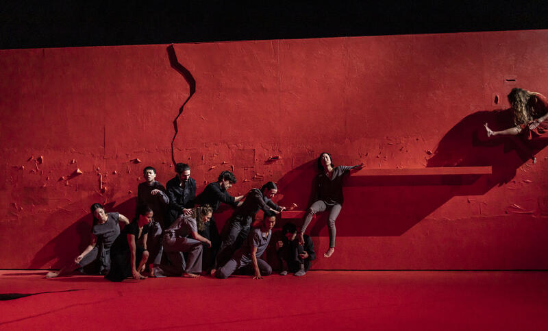 Flera dansare står mot en röd vägg som krackilerar, en dansare är uppfäst på den. Dansarna är fint upplysta som gör att bilden ser konstnärlig ut på den scen de står 