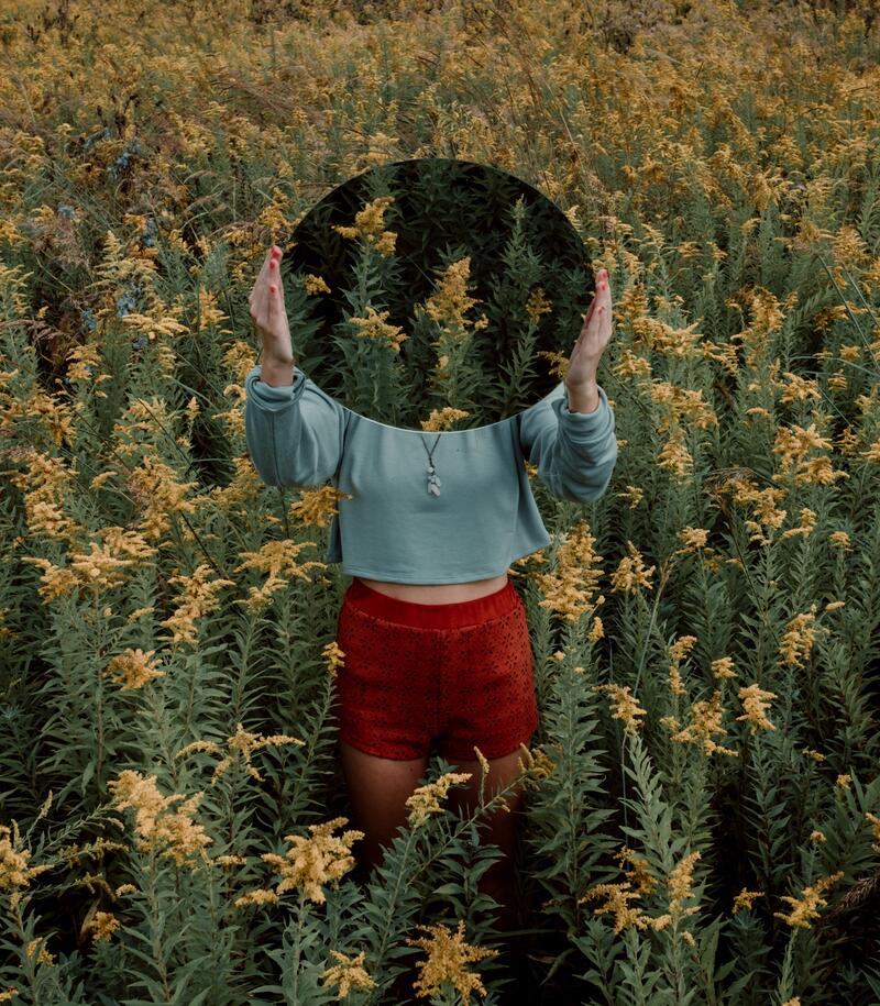 Woman in field holding a mirror covering here face and replacing it for a view of the field. The field is yellow and quite tall. 