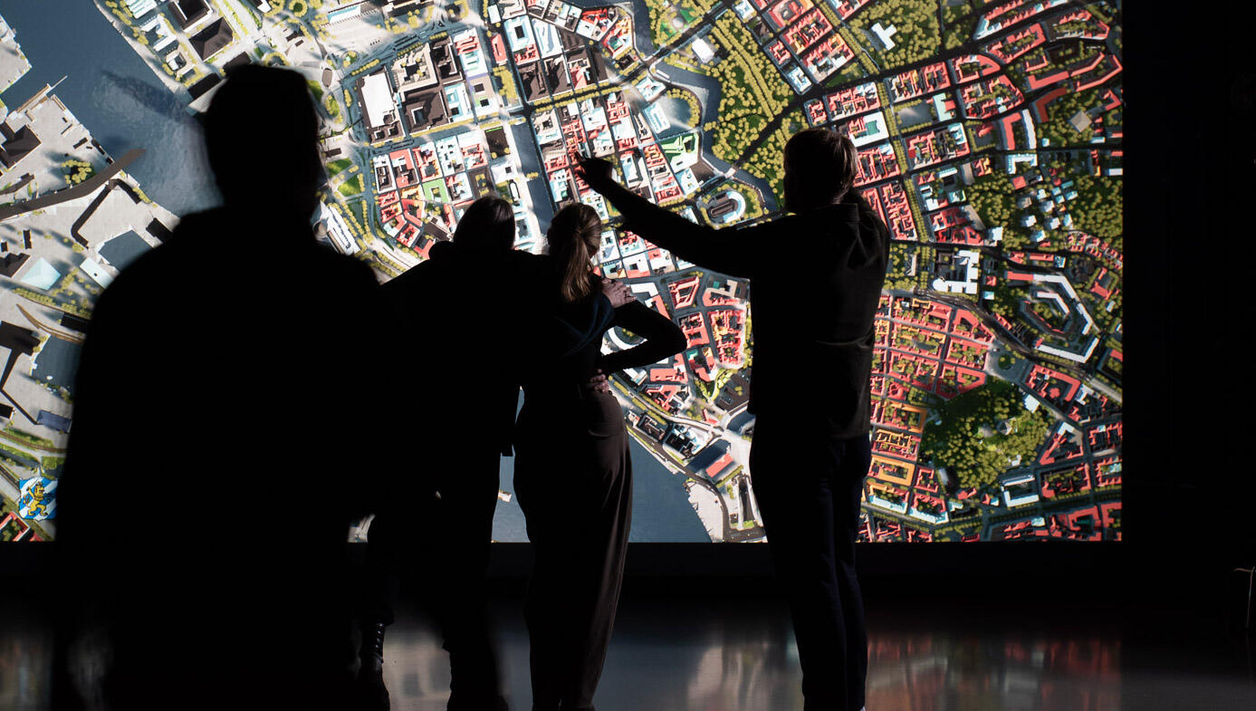 Tre människor står och pekar mot en stor skärm där ett flygfoto av en stad projiceras. 
