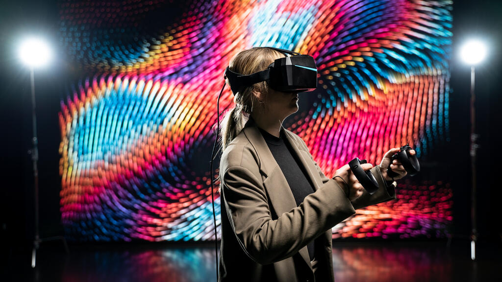 Kvinna labborerar i VR. Bakgrunden är futuristisk och färglad och föreställer digitala vågor som rör sig. Kvinnan är coolt upplyst och står i en brun kostym. Två bakgrundslampor skymtas i öven i rummet som i övrigt rummet är mörkt belyst. 