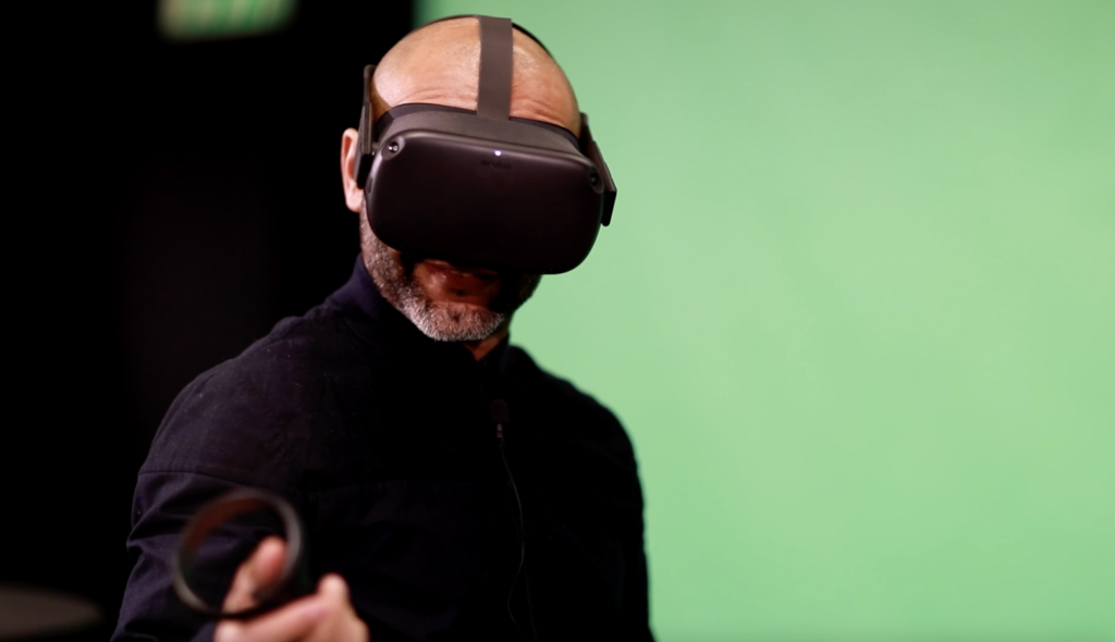 En man som står med ett VR-headset. Som ett par glasögon fast hela hans omgivning är virtuell när han tittar genom glasögonen. I fotot ser man bara mannen som ser glad ut . Bakgrunden är grön. 