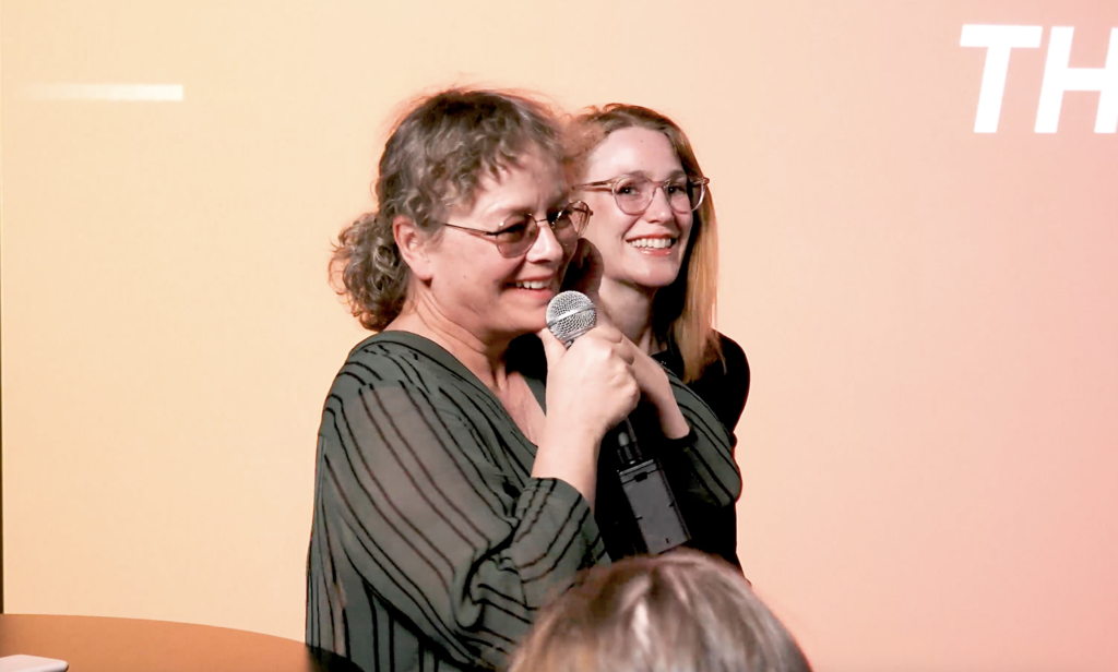 Två kvinnor på en scen, leendes utöver publiken. 