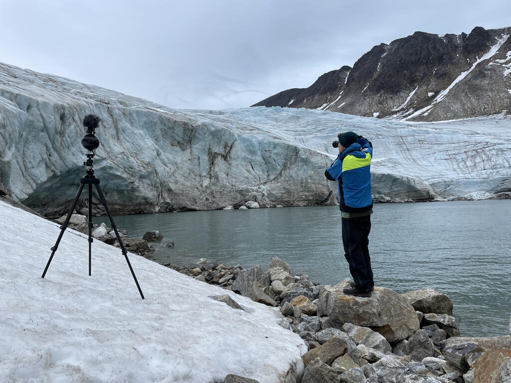 En man (Forksaren Tyrone Martinsson) vid en vattenkant som fotograferar en glaciär 