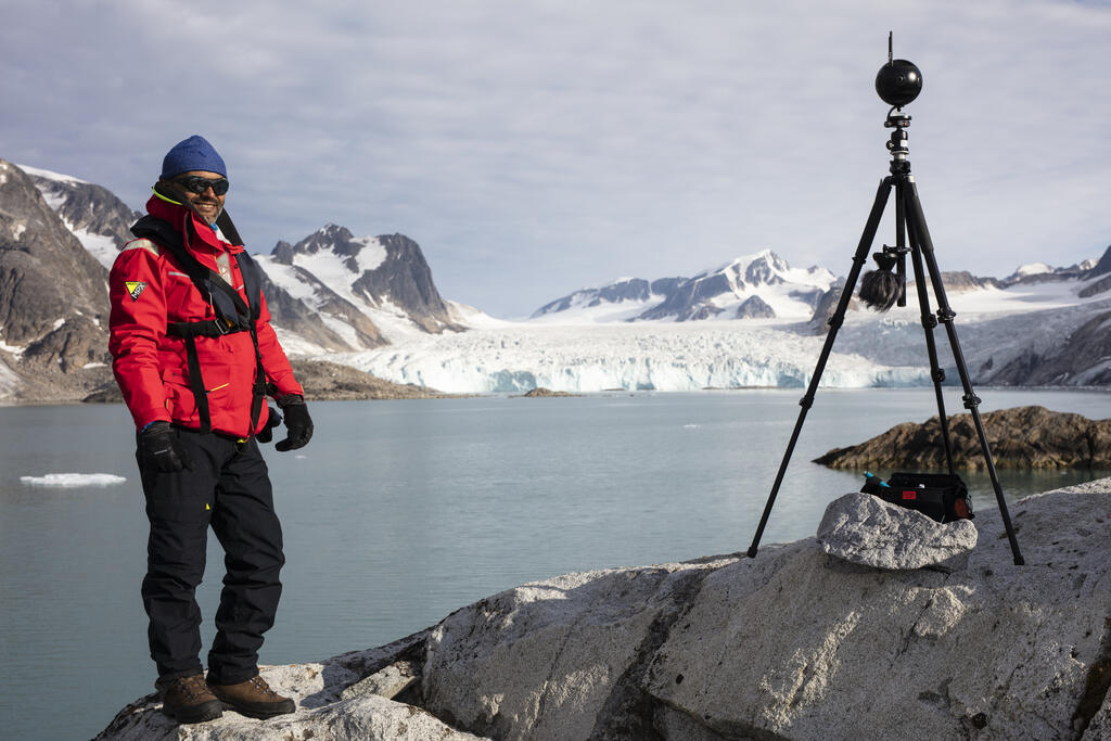 Man på Svalbard, ler, solsken, mannen står på en klippa i bakgrunden öppnar en sjö upp sig som sedan går över i en glaciär 