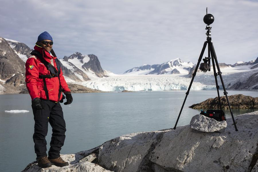 Man står med expeditionskläder på svalbard, i bakgrunden öppnar en stor glaciär sig upp. Bredvid mannen syns en 360 graders kamera på ett kamera stativ! 