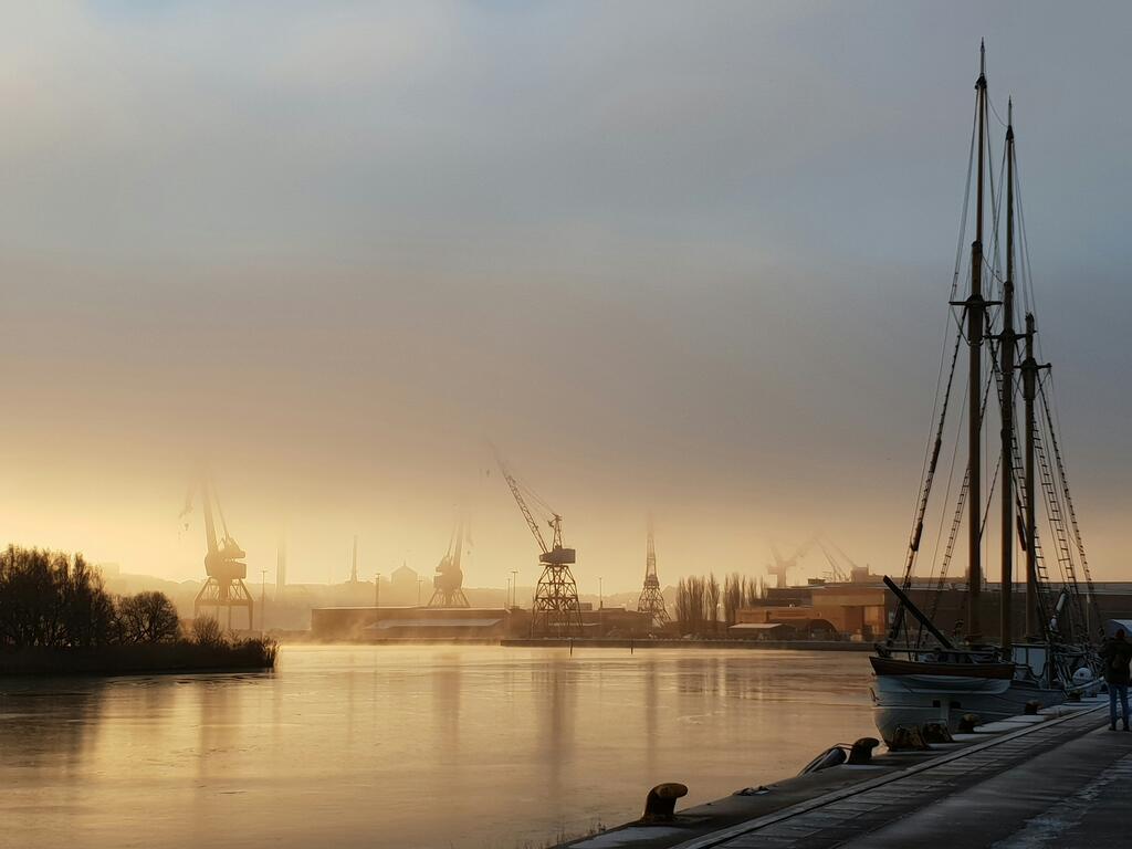 Göteborg en soldisig morgon, gamla varvskranar syns mot horisonten med en lungt vatten i förgrunden. 